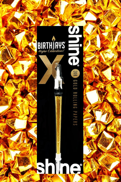 Black packaging for “Shine 24K Gold Rolling Papers” featuring a cannabis oil applicator, displayed against a backdrop of gold foil-wrapped chocolate squares.