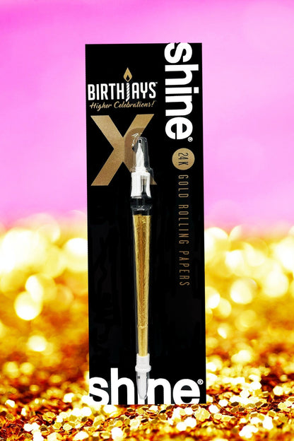 A black and gold package of Shine 24K gold rolling papers with a pre-rolled joint visible, set against a blurred golden confetti background.