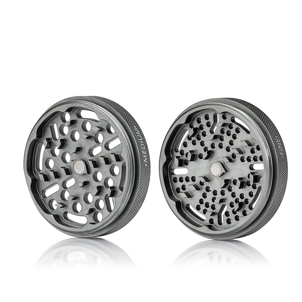 Front-facing studio shot of two metallic RYOT GR8TR Solid Body Grinder halves with textured edges and patterned grinding holes reflected on a white surface