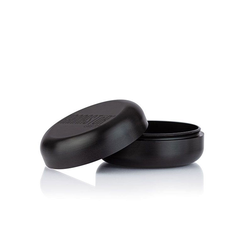 Three-quarter view of the matte black RYOT GR8TR Solid Body Grinder with its lid tilted beside the base on a reflective white surface
