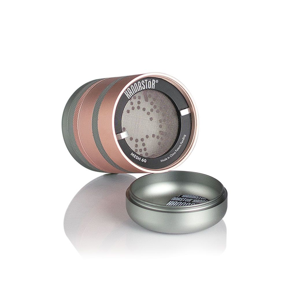 Angled close-up of the RYOT GR8TR Solid Body Grinder showing its rose-gold and gunmetal stacked chambers and a detached lid on a reflective white surface