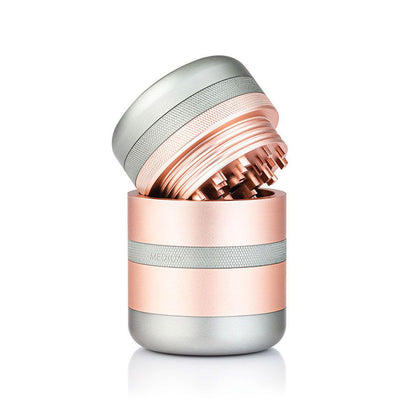 Angled close-up of the metallic rose gold and silver RYOT GR8TR Solid Body Grinder with its lid tilted to reveal interlocking teeth and knurled grip bands.