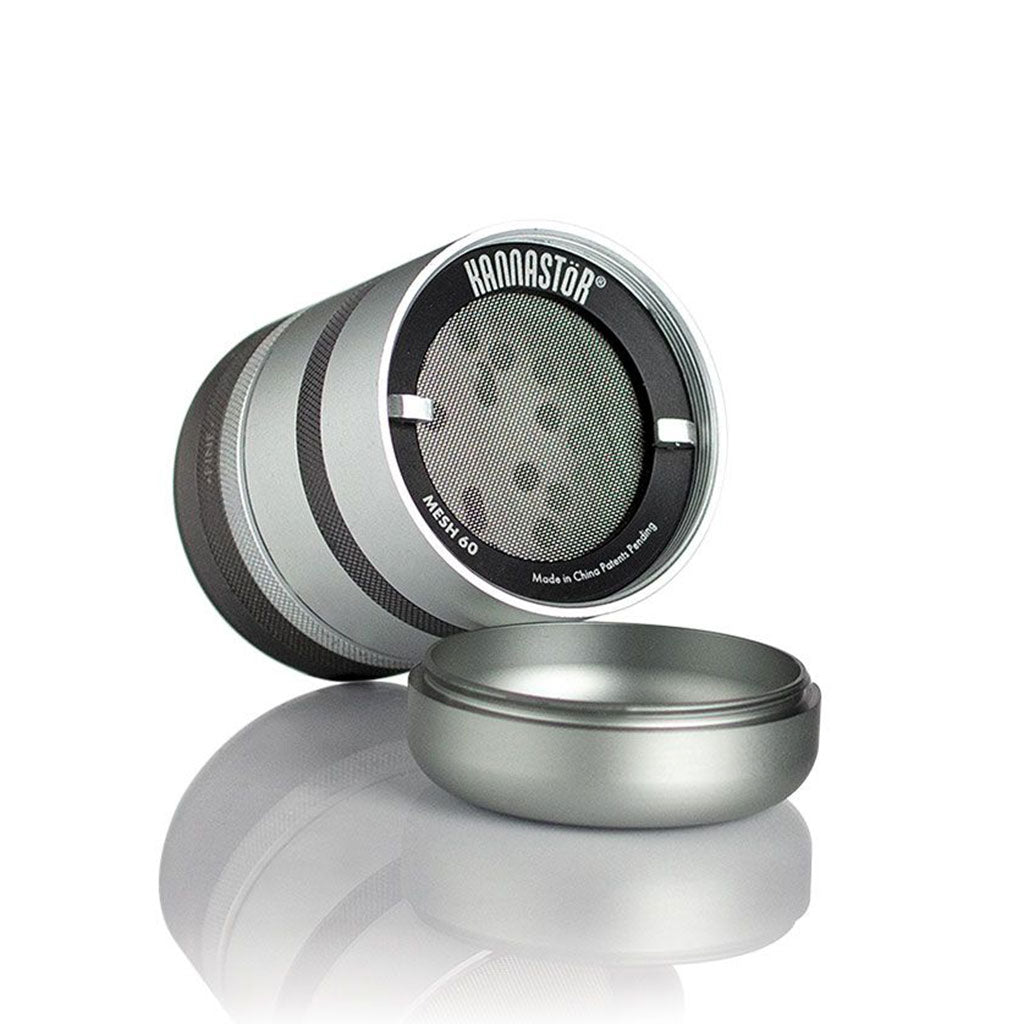 Angled studio shot of the metallic RYOT GR8TR Solid Body Grinder showing its open chamber, fine mesh screen, and soft reflection on a white surface