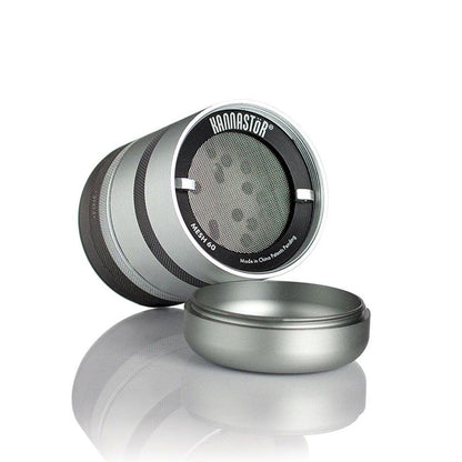 Angled studio shot of the metallic RYOT GR8TR Solid Body Grinder showing its open chamber, fine mesh screen, and soft reflection on a white surface