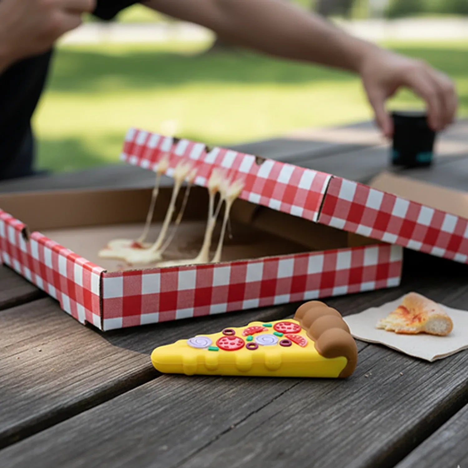 Close-up on a yellow Cloud 8 Silicone Pizza Slice Hand Pipe shaped like a pepperoni pizza slice resting on a picnic table in front of a red-and-white checkered pizza box.