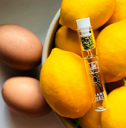 A glass vial filled with cannabis buds stands among a pile of ripe mangoes, branded with ""Jane West"" and a sun logo."