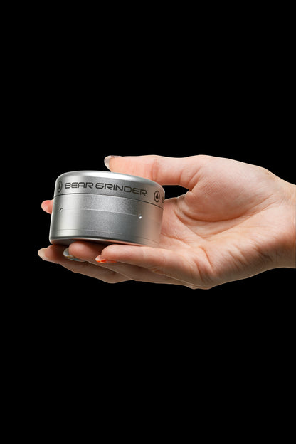 A hand holding a sleek, metallic cannabis grinder labeled “Bear Grinder” against a black background.