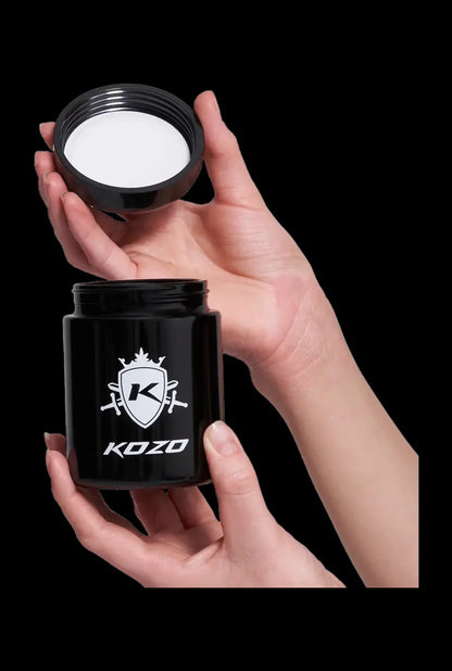 A hand holds a clear Kozo Glass jars - 1/2 OZ jar, catching the light with a smooth, rounded design.