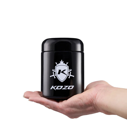 Handheld front view of a glossy black Kozo Glass jars - 1/2 OZ with white crest logo centered on the jar.