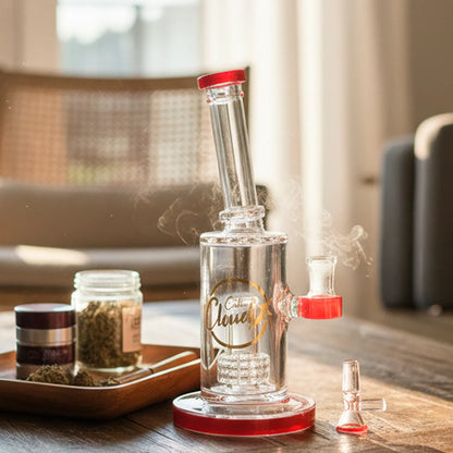 Angled close-up of the Smoke Cartel 8-inch Showerhead Dual-Use Water Pipe w/ Colored Accents showing clear glass with red accents, light smoke, and jars on a wooden table