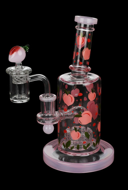 Angled front view of the Pulsar Fruit Series Peaches & Cream Glow in The Dark Dab Rig Kit - 8" / 14mm F showing pink peach decals on a clear glass body with a matching pink base and detached peach-topped banger.