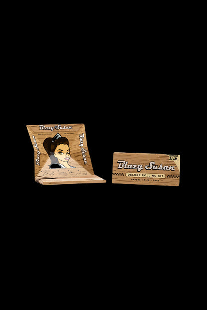 A product shot of a ""Blazy Susan"" deluxe rolling kit, featuring slim rolling papers, tips, and a small tray, with vintage-style branding and a portrait of a woman on the packaging."