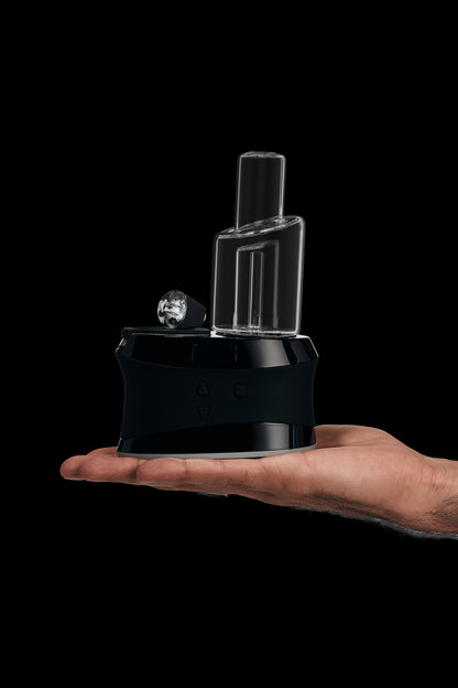 A hand holds a sleek, black cannabis e-rig with a glass water pipe attachment against a dark background.