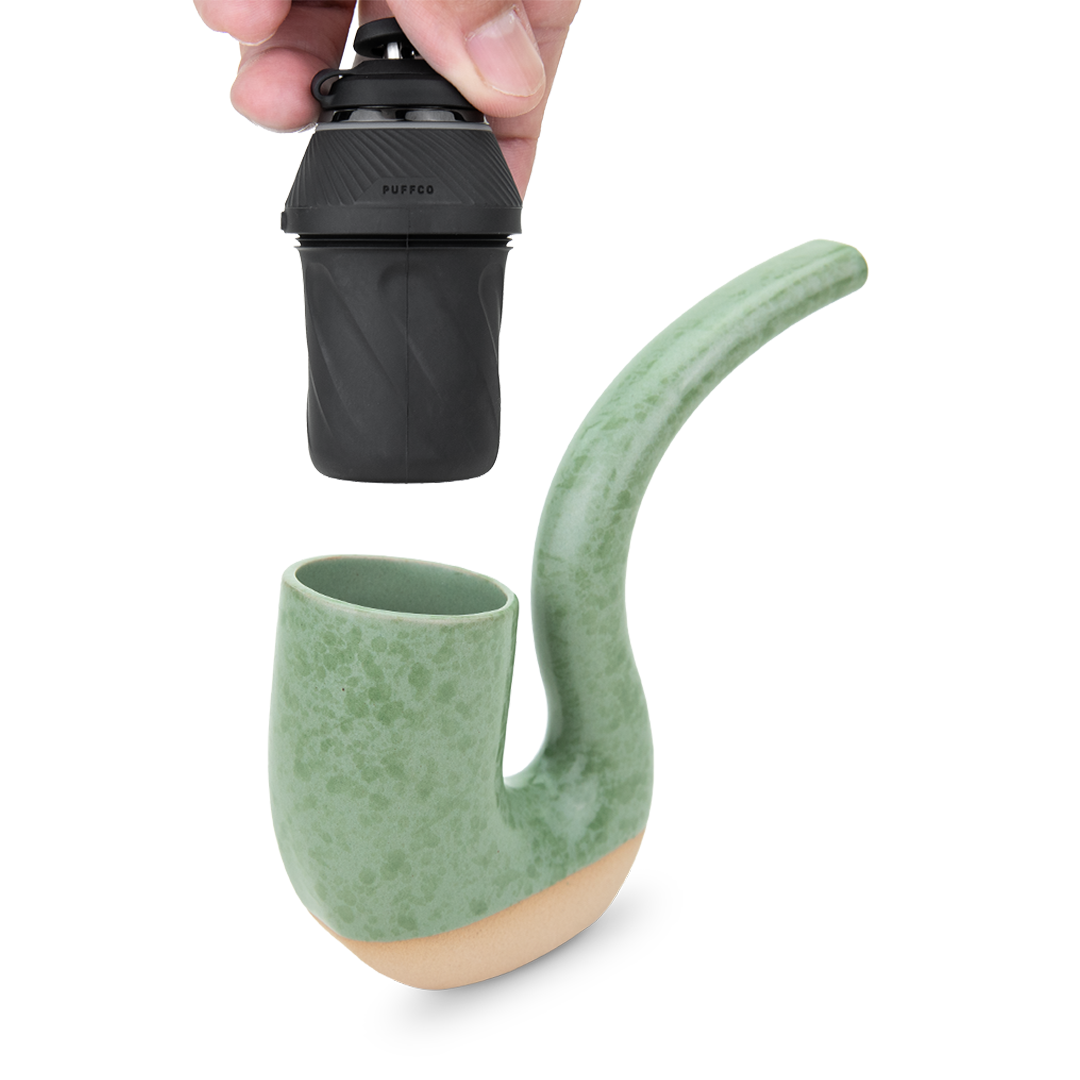 Top-down view of a hand holding the black Puffco Proxy Terrapipe mouthpiece above a speckled green and tan ceramic pipe body on a white background