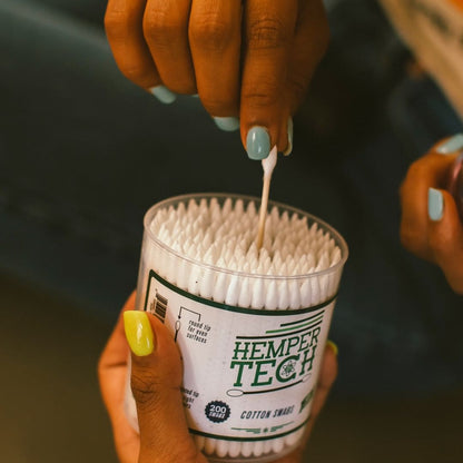Close-up of a hand pulling a cotton swab from a clear tub of Hemper Tech Cotton Buds 200ct, showing packed white tips and a dark blurred background