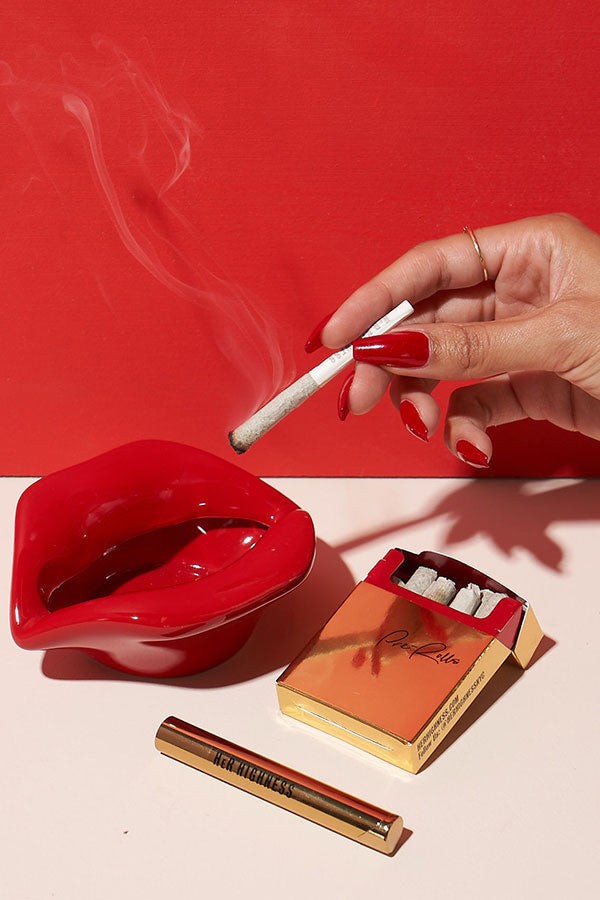 A hand with red nails holds a lit joint above an ashtray and matches, all set against a red and pink background. 