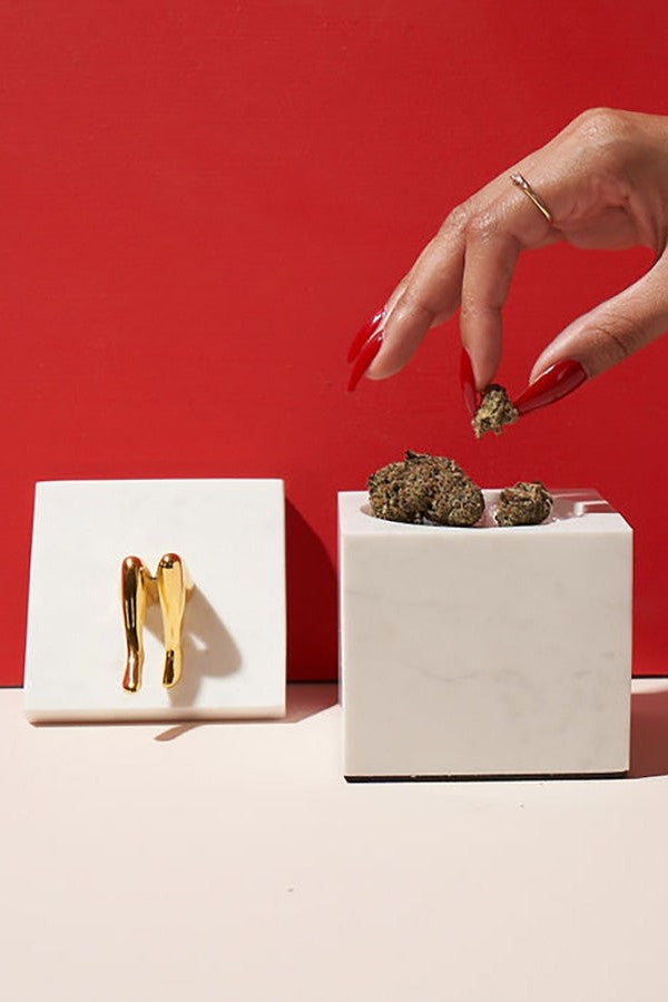 Hand with red nails placing cannabis bud into a white container alongside golden tweezers, set against a red background.