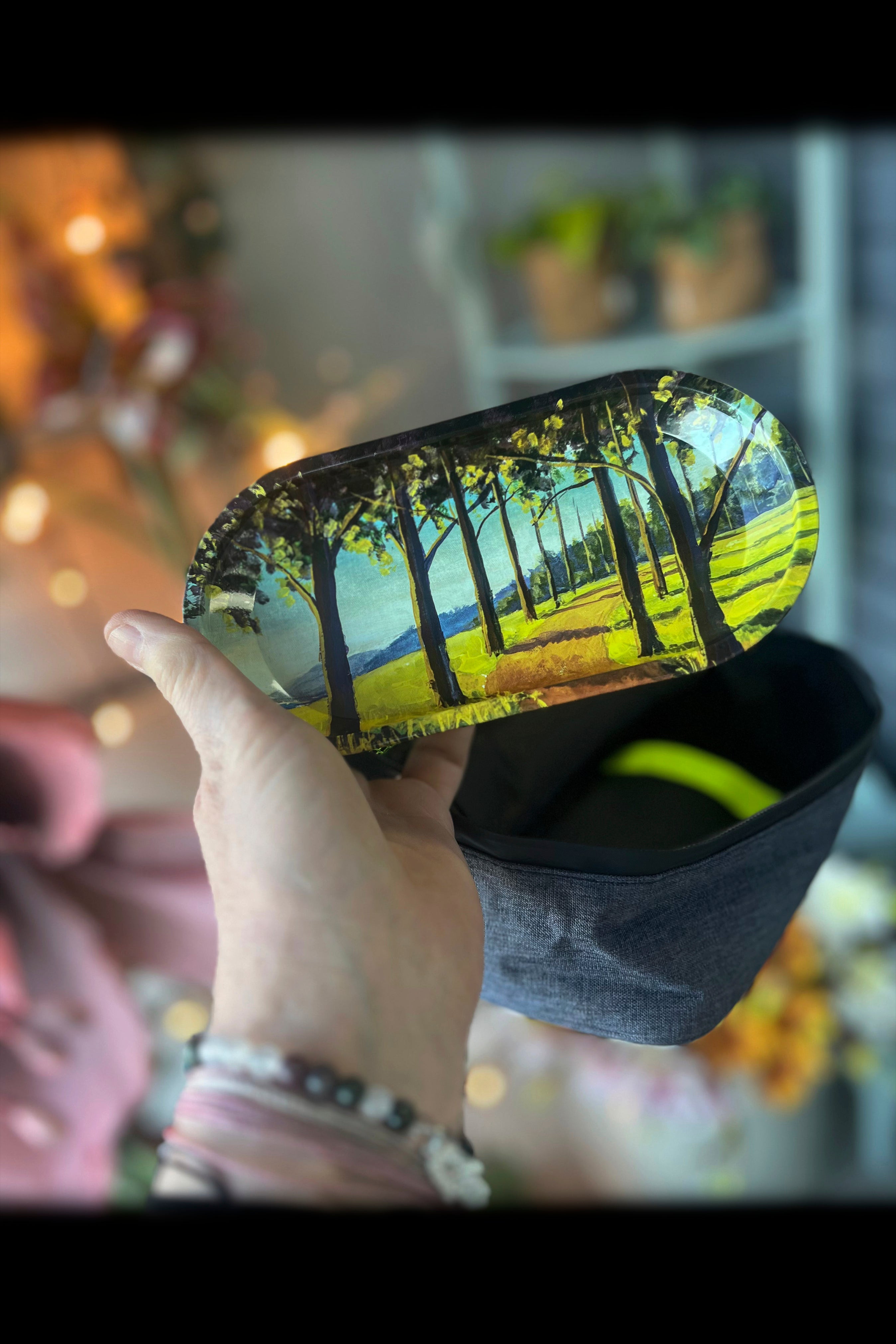 Hand holding a colorful, painted rolling tray depicting a forest scene, above a dark blue stash bag with a green cannabis leaf visible inside.