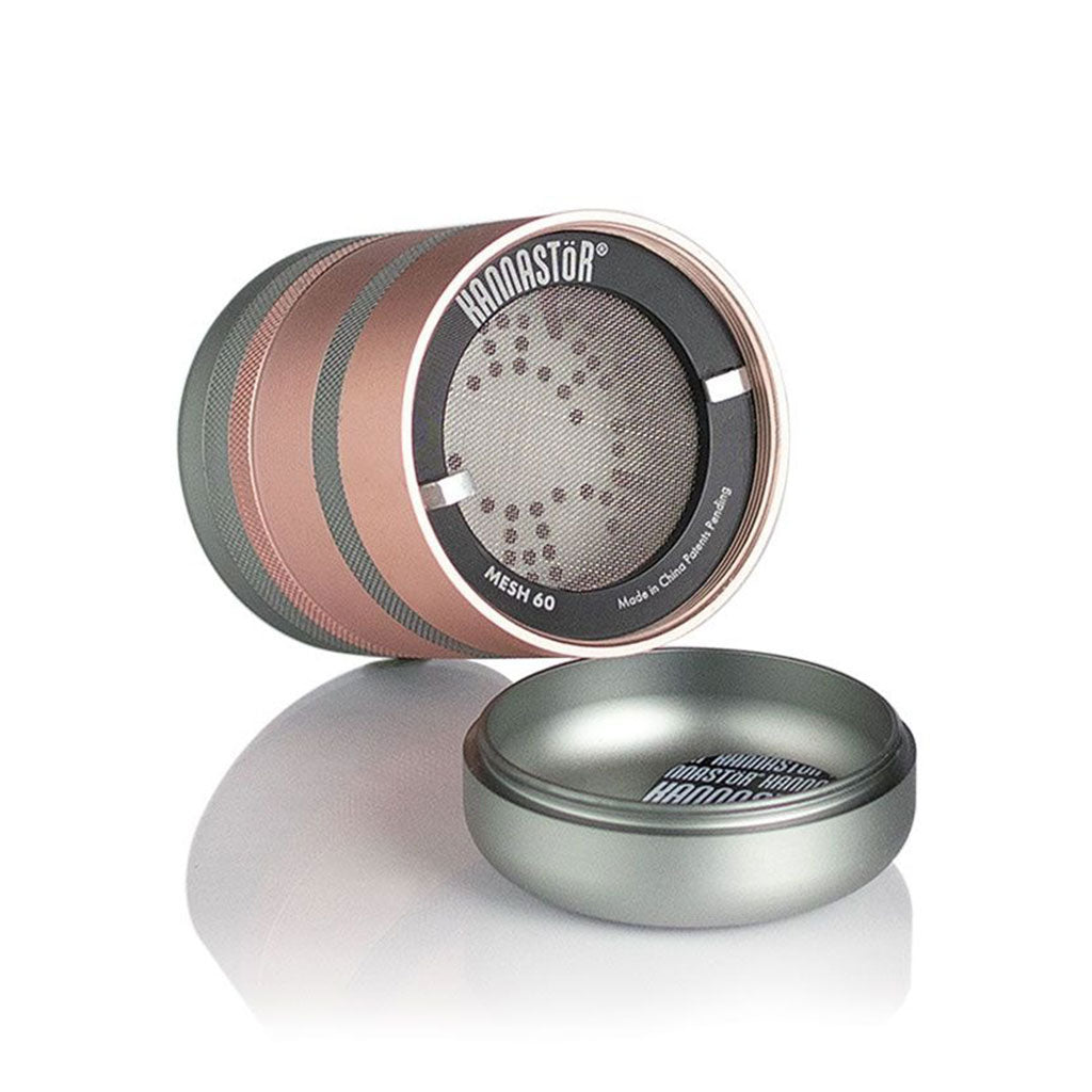 Angled close-up of the rose-gold and silver RYOT GR8TR Jar Body Grinder showing its mesh screen and separated lid reflected on a white surface