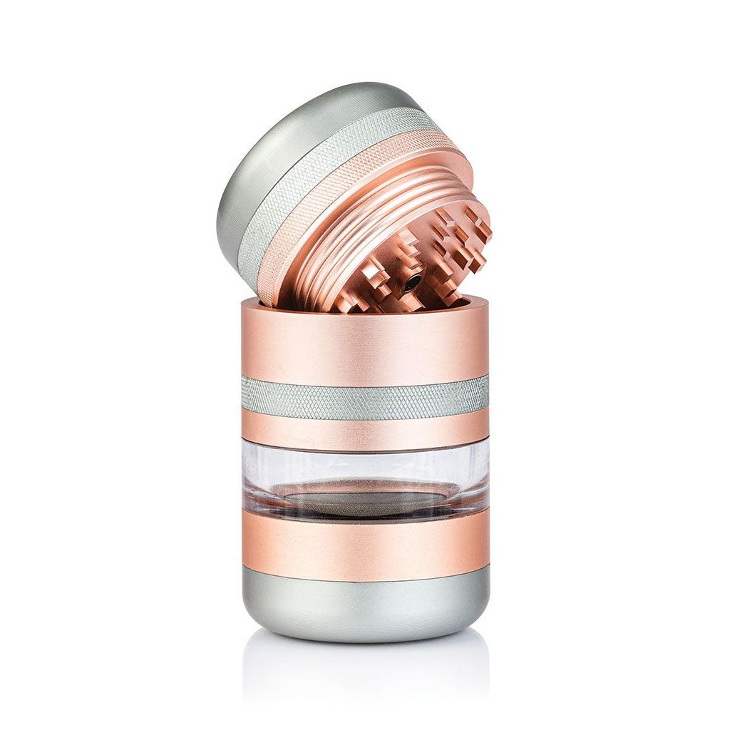 Angled close-up of a rose-gold and silver RYOT GR8TR Jar Body Grinder with its toothed lid tilted to show internal teeth and clear middle chamber