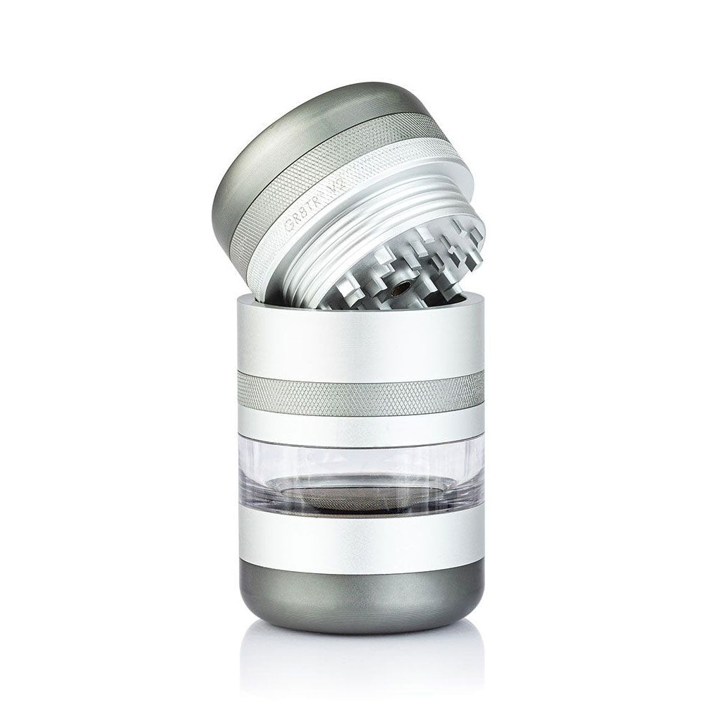 Angled close-up of the silver RYOT GR8TR Jar Body Grinder with its lid tilted to show sharp internal teeth and a clear middle chamber.