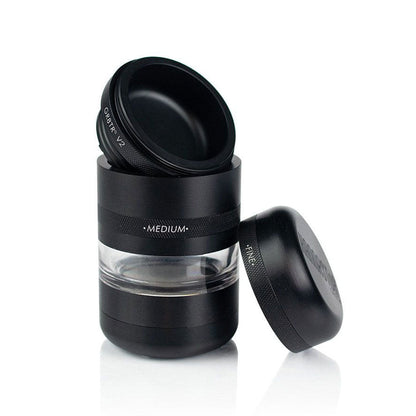 Three-quarter view of the black RYOT GR8TR Jar Body Grinder with its lid removed and propped beside the clear middle chamber.