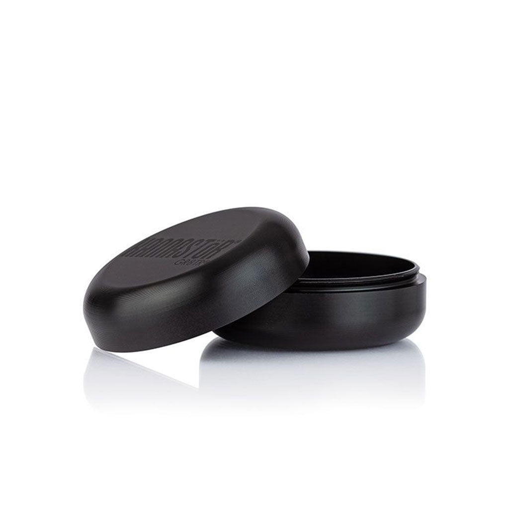 Angled close-up of the black RYOT GR8TR Jar Body Grinder with its lid propped beside the open base on a white reflective surface