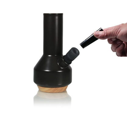 Angled front view of the black RYOT Ceramic Maple Traveller Pipe on a maple base with a hand holding a detachable mouthpiece against a white background
