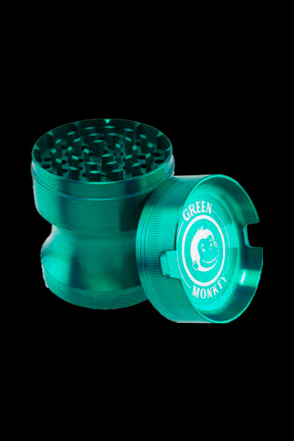 Angled close-up of a metallic Green Monkey Chacma Grinder with Ashtray in black-green lighting showing its toothed grinding chamber and branded ashtray lid.