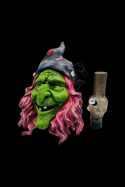 Green witch mask with pink hair alongside a glass bubbler pipe on a black background.