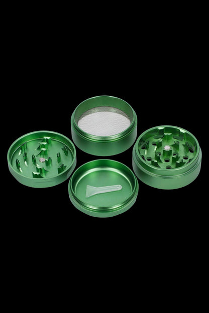 Green four-piece cannabis grinder disassembled, showing the lid, grinding chamber, filter screen, and collection chamber with a small scraping tool.