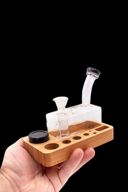 A hand holds a wooden dab station with a glass oil rig and accessories against a black background.