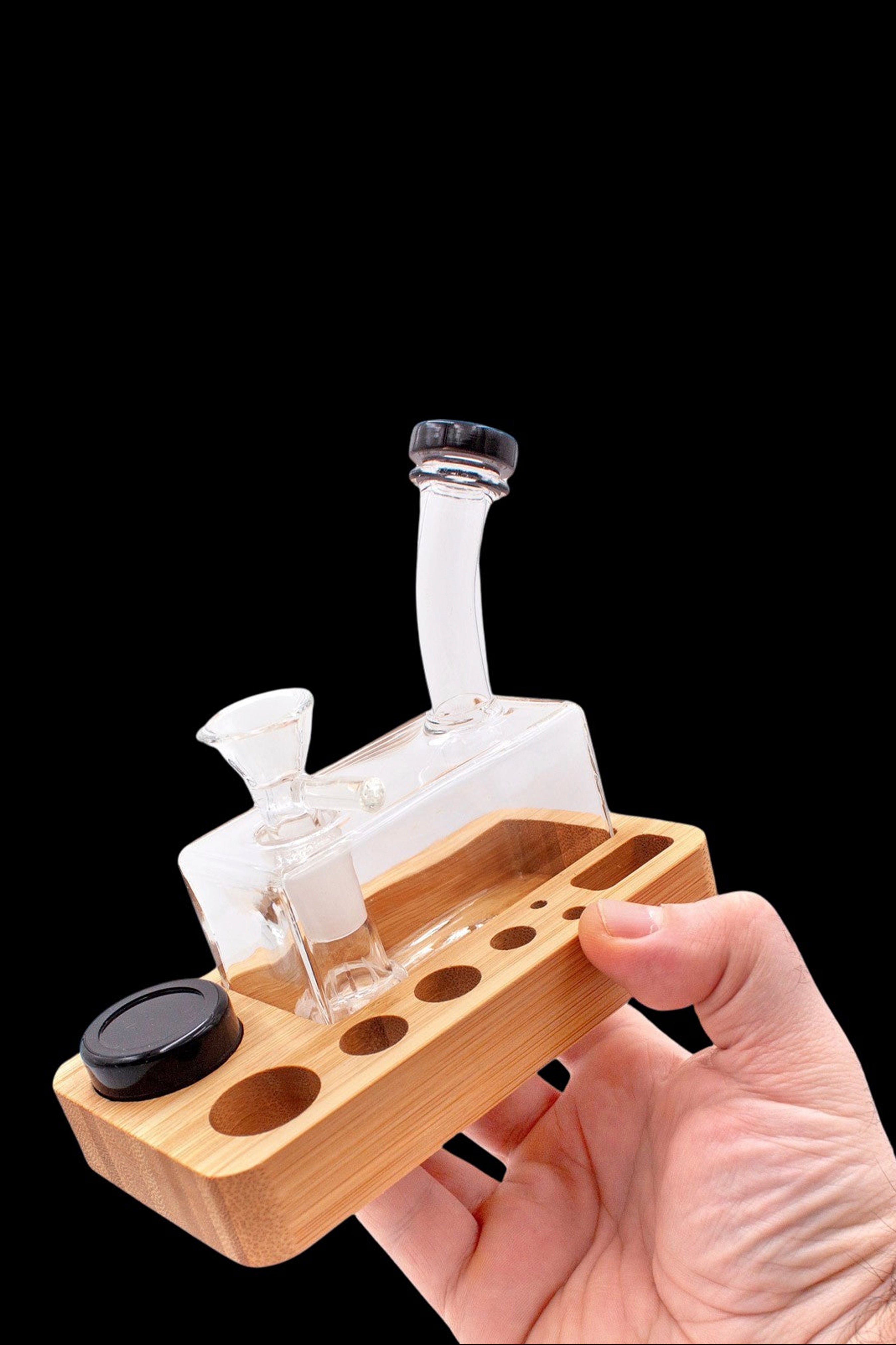 A hand holds a wooden rolling tray with a small glass water pipe (bong) and containers for cannabis concentrates.