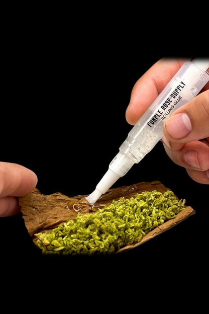 Close-up of a hand applying Purple Rose Supply Cannagar & Blunt Rolling Glue from a clear applicator tip onto a brown leaf filled with green herb against a black background