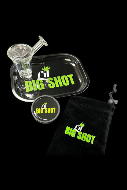 A glass bubbler pipe with a “Big Shot” branded sticker and black drawstring bag, set against a dark background.
