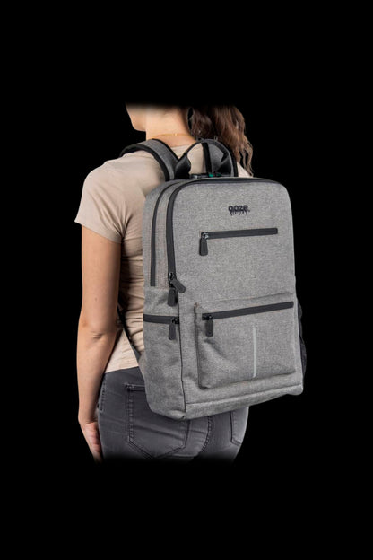 Rear three-quarter view of a woman wearing the Ooze Traveler Smell Proof Backpack in light gray against a black background.