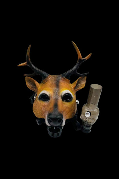 A deer mask with glowing eyes holds a glass cannabis oil rig in its mouth against a black background.