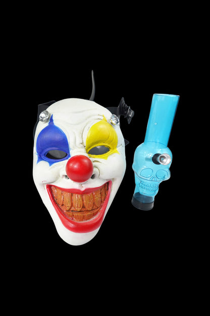 Creepy clown mask with a blue glass pipe positioned beside it on a black background.