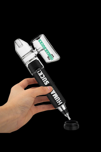 Hand holding a clear and black cannabis oil rig with the words "HUMAN SUCK" printed on the side, above a small collection cap on a dark background.