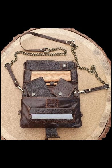 Brown leather cannabis accessory pouch with multiple pockets and a chain strap, displayed on a wood slice.