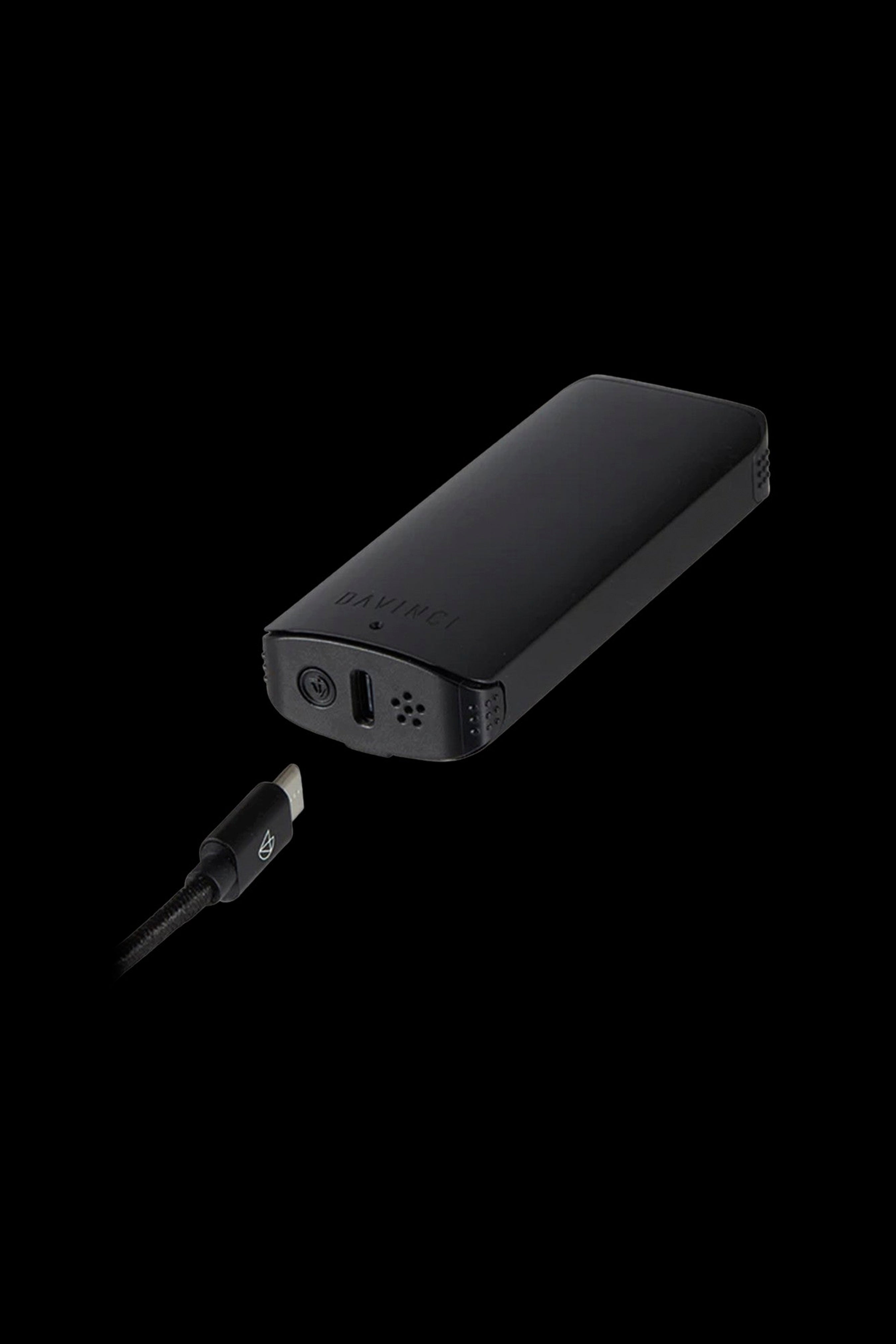 Black Davinci IQ2 vaporizer with a charging cable plugged in, isolated on a black background.