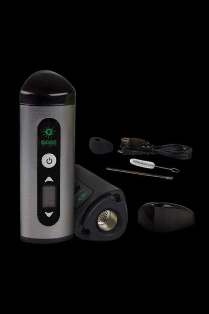 Silver cannabis vaporizer pen with included mouthpiece, cleaning tools, and charging base displayed on a black background.