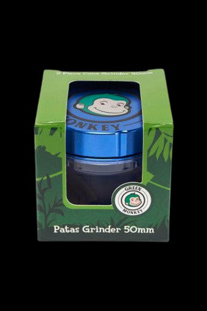 Top-front view of the Green Monkey Patas Grinder visible through its green windowed box, showing the blue metal lid with the monkey logo.