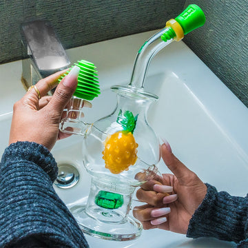 Hands assembling a glass water pipe (bong) shaped like a pineapple, with green accents and cannabis bud visible inside. - Hemper Tech Cleaning Plugs + Caps