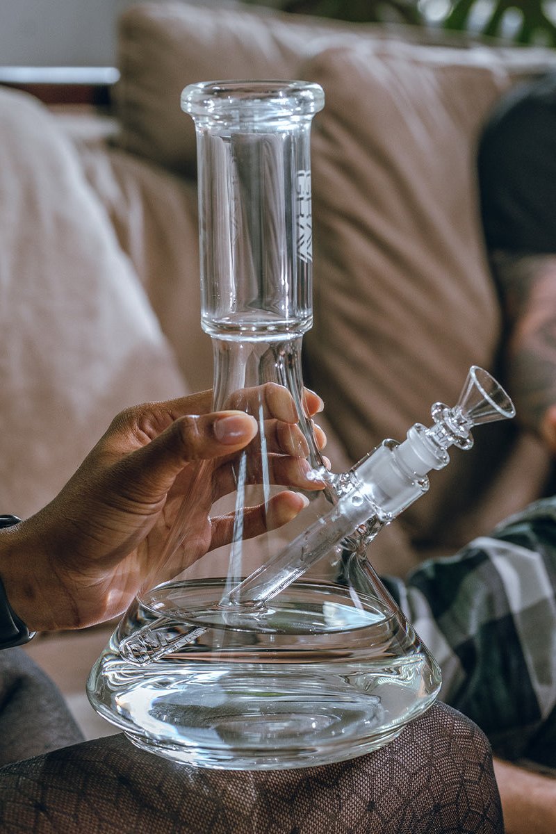 A person holds a clear glass water pipe (bong) with a detachable bowl piece, likely for cannabis use, against a blurred background of a beige couch.