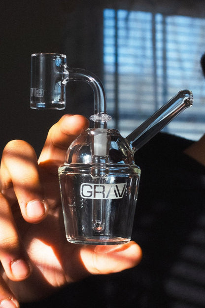 A hand holds a clear glass dab rig with the brand “GRAV” printed on the side, with a separate glass carb cap blurred in the background.