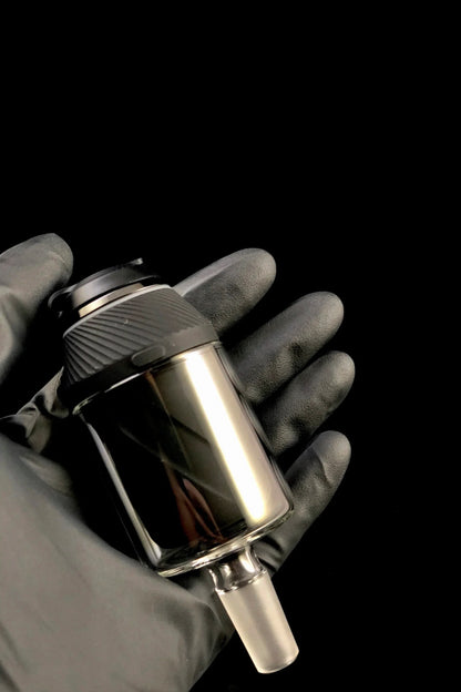 A gloved hand holds a sleek, metallic cannabis oil rig with a glass chamber.