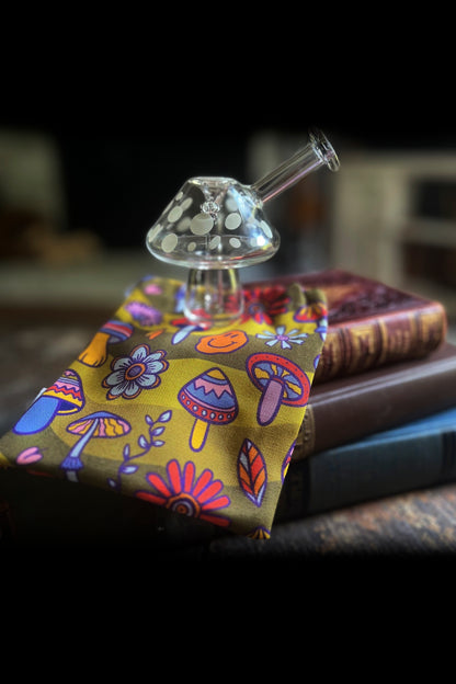 A glass mushroom pipe rests on a patterned bandana draped over stacked vintage books, illuminated in a dimly lit, moody setting.