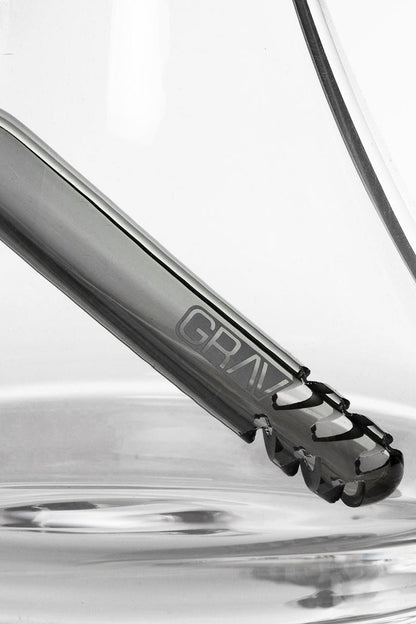 Close-up of a glass cannabis water pipe with the brand name "GRAV" visible, partially submerged in rippling water.