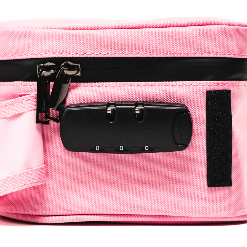 Close-up side view of a pink Blazy Susan Stash Bag showing black zipper pulls and an attached three-digit combination lock against textured fabric.